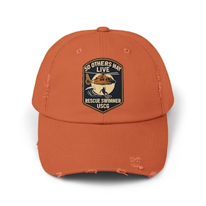 May include: Burnt orange distressed baseball cap featuring a patch with a helicopter, a swimmer, and the text "SO OTHERS MAY LIVE RESCUE SWIMMER USCG". The cap has a curved brim and a worn appearance.