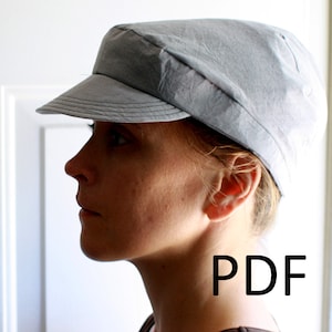 Könnte beinhalten: Eine Frau trägt eine graue, flache Kappe mit einem braunen Shirt. Das Wort "PDF" ist in Schwarz auf dem Shirt aufgedruckt.