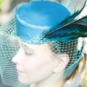 May include: A teal-colored hat with a cylindrical crown, adorned with dark teal and black feathers. A matching veil drapes over the face. The word "PDF" is printed in black at the top of the image. This headwear is suitable for special occasions.