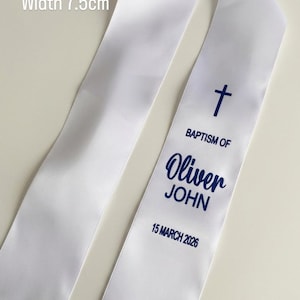 May include: A white satin sash measuring 90 cm in length and 7.5 cm in width, featuring a dark blue cross, the text BAPTISM OF Oliver JOHN, and the date 15 MARCH 2026. The ends of the sash are finished with a delicate white lace trim detail.