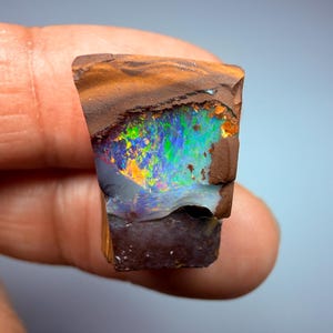 May include: A raw boulder opal specimen featuring a dark brown ironstone matrix with a vibrant, iridescent play-of-color in shades of neon green, electric blue, and fiery orange. The gemstone displays a natural, irregular shape with a polished face.