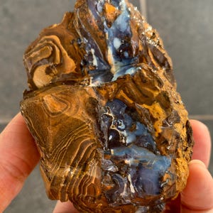 Puede incluir: Un espécimen de ópalo boulder en bruto que presenta una veta central de ópalo azul y blanco iridiscente incrustada en una matriz texturizada y estratificada de piedra de hierro marrón y beige con bandas concéntricas y acabado brillante.