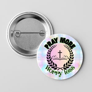 May include: A round button with the text "PRAY MORE WORRY LESS" in green, surrounding a black cross and open book design. The background is a pastel plaid pattern. The back of the button is silver.