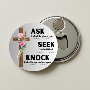 Puede incluir: Un abrebotellas redondo con acabado metálico plateado. El abridor presenta un diseño circular blanco con una cruz de madera adornada con flores rosas y el texto "ASK, SEEK, KNOCK".