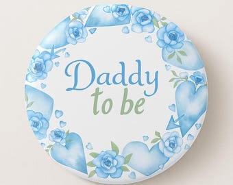 May include: A round, white button with the text "Daddy to be" in blue and green script. The button is decorated with a wreath of blue watercolor roses, hearts, and arrows.
