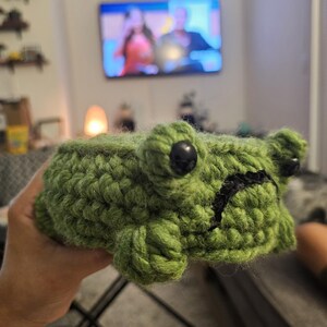 Puede incluir: Una rana verde de ganchillo hecha a mano, con ojos negros y boca negra. La rana est&aacute; hecha de hilo texturizado y se sostiene en una mano. Un art&iacute;culo decorativo hecho a mano.