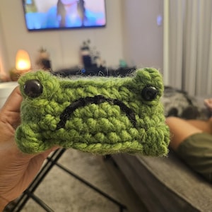 Puede incluir: Una funda hecha a mano con forma de rana de ganchillo verde con ojos de botón negros y una boca cosida en negro. La funda está sujeta en la mano de una persona.