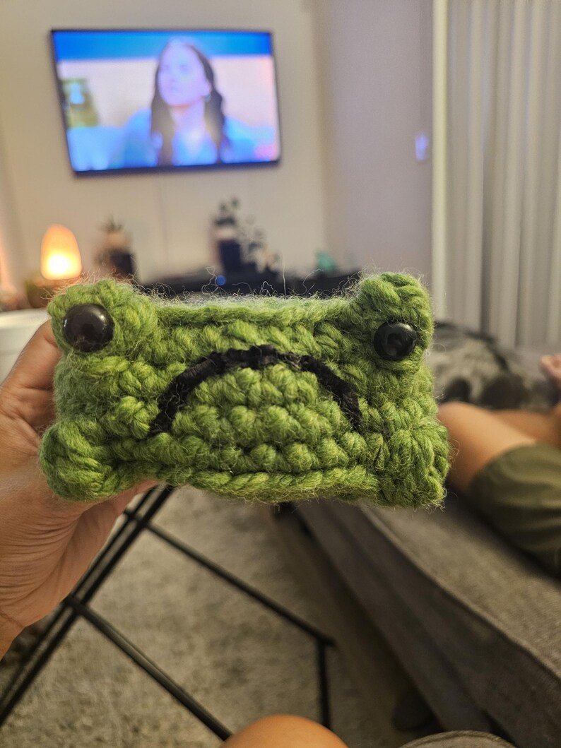 Puede incluir: Una funda hecha a mano con forma de rana de ganchillo verde con ojos de bot&oacute;n negros y una boca cosida en negro. La funda est&aacute; sujeta en la mano de una persona.