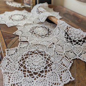 May include: Cream-coloured, handmade crochet doilies and a table runner. The doilies have a circular, openwork design with scalloped edges. The table runner is made of the same material and design, with a long, rectangular shape. The items are displayed on a wooden surface.
