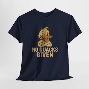 以下が含まれることがあります： ネイビーブルーのTシャツ。アヒルがウインクし、腕を組んだ漫画のイラストが描かれています。「NO QUACKS GIVEN」の文字がアヒルのイラストの下に金色でプリントされています。カジュアルでユーモラスなデザインです。