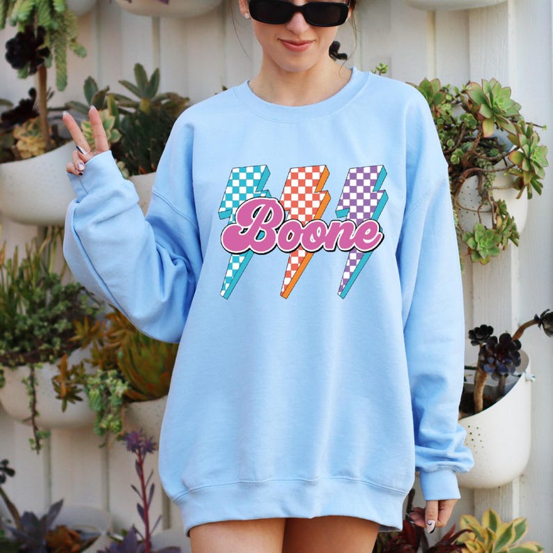 May include: A light blue sweatshirt with the name "Boone" in pink script, overlaid on three lightning bolt graphics. The lightning bolts have checkered patterns in teal, orange, and purple. The sweatshirt is a solid light blue color.