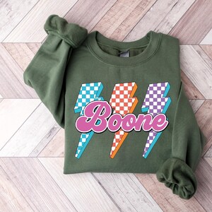 May include: Olive green sweatshirt with a graphic design featuring three lightning bolts in blue, orange, and purple checkered patterns. The word "Boone" is written in pink script across the lightning bolts.