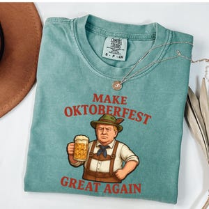 May include: A teal Comfort Colors t-shirt with the words "MAKE OKTOBERFEST GREAT AGAIN" in red. The graphic depicts a cartoon figure in traditional Bavarian attire holding a beer stein. A silver necklace with a pendant is also visible.