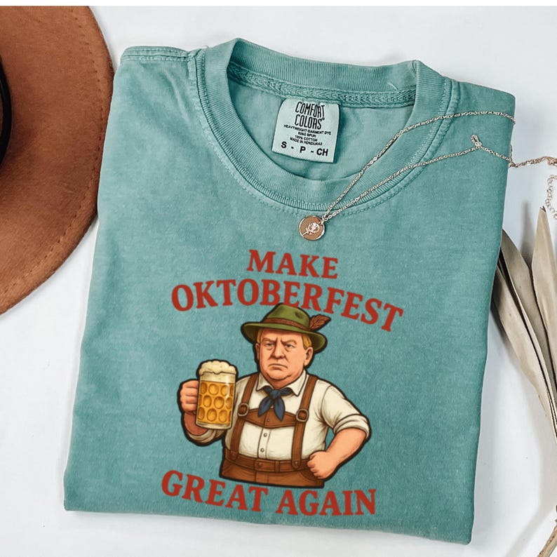 May include: A teal Comfort Colors t-shirt with the words "MAKE OKTOBERFEST GREAT AGAIN" in red. The graphic depicts a cartoon figure in traditional Bavarian attire holding a beer stein. A silver necklace with a pendant is also visible.