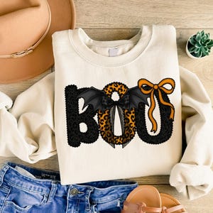May include: Cream-colored sweatshirt with the word "BOO" in a Halloween-themed design. The "B" is black with a textured pattern, the "O" is leopard print with bat wings, and the second "O" has an orange bow.