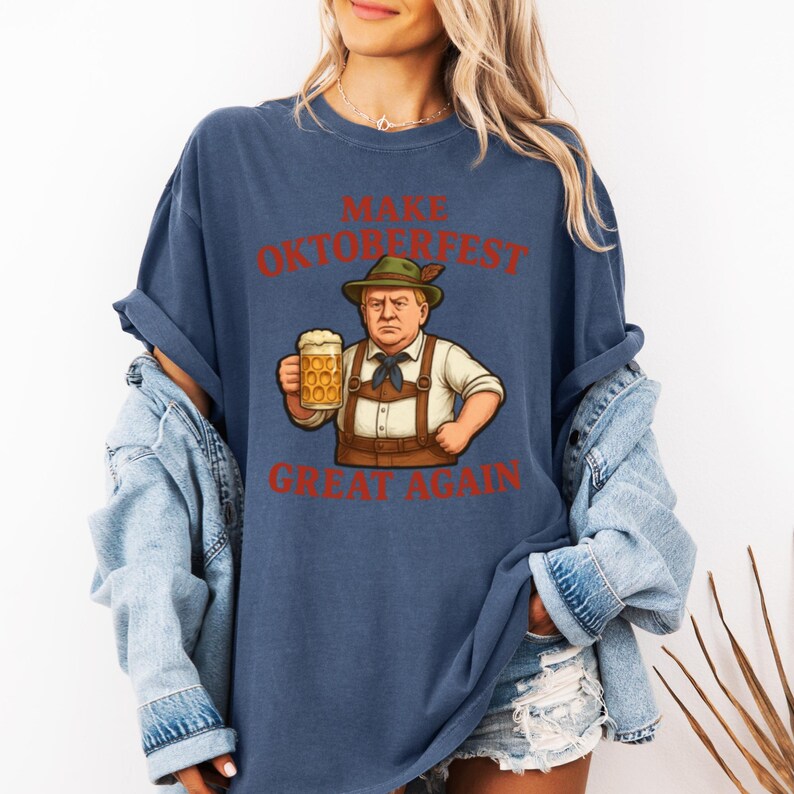 May include: Navy blue t-shirt with the text "MAKE OKTOBERFEST GREAT AGAIN" in red. The graphic features a cartoon of a man in traditional Bavarian attire holding a beer stein. The shirt is a casual, oversized fit.