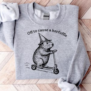 May include: A heather gray sweatshirt featuring a black and white illustration of a beaver wearing a witch's hat and riding a scooter. The text "Off to cause a kerfuffle" is printed above the beaver.