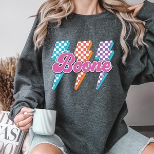 May include: Dark gray sweatshirt with the name "Boone" in a pink, retro font, with three lightning bolt graphics in blue, orange, and purple checkered patterns. The sweatshirt is a casual style.