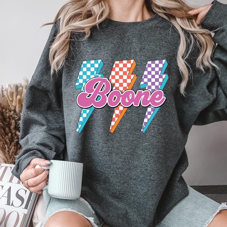 May include: Dark gray sweatshirt with the name "Boone" in a pink, retro font, with three lightning bolt graphics in blue, orange, and purple checkered patterns. The sweatshirt is a casual style.