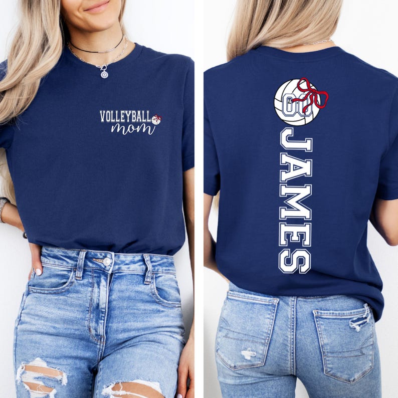 May include: Navy blue t-shirt with the text "Volleyball Mom" in white script on the front. The back of the shirt features a volleyball graphic with a red bow and the name "JAMES" in white vertical lettering.