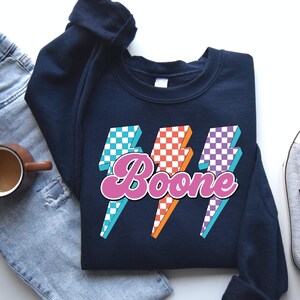 May include: Navy blue sweatshirt with a retro design featuring three lightning bolts in blue, orange, and purple checkered patterns. The word "Boone" is written in pink script across the center of the design.