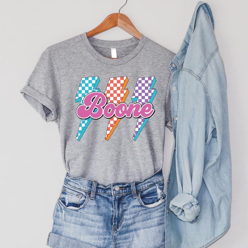 May include: A gray t-shirt with the name "Boone" in a retro font, set against three lightning bolt graphics. The lightning bolts feature checkered patterns in teal, orange, and purple. The shirt is paired with denim shorts and a denim jacket.