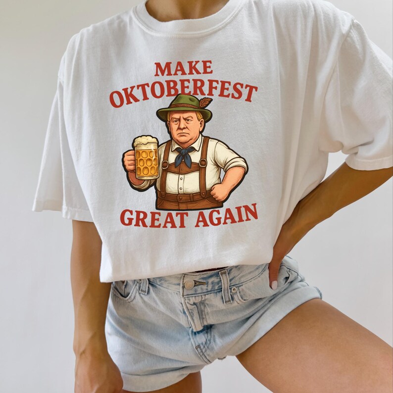 May include: White t-shirt with the text "MAKE OKTOBERFEST GREAT AGAIN" in red. The shirt features an illustration of a man in traditional Bavarian attire holding a beer stein. The design is centered on the front of the shirt.
