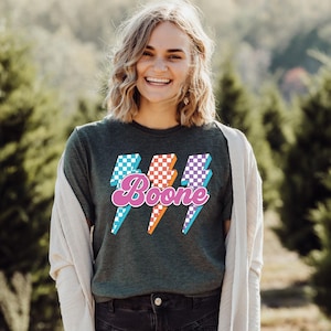 May include: Dark green t-shirt with the name "Boone" in pink script, overlaid on three lightning bolt graphics. The lightning bolts have checkered patterns in blue, orange, and purple. The person is wearing a light-colored cardigan.