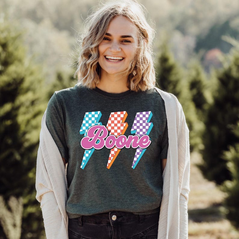 May include: Dark green t-shirt with the name "Boone" in pink script, overlaid on three lightning bolt graphics. The lightning bolts have checkered patterns in blue, orange, and purple. The person is wearing a light-colored cardigan.