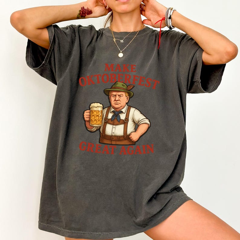 May include: Dark grey t-shirt with the text "MAKE OKTOBERFEST GREAT AGAIN" in red. The shirt features a cartoon illustration of a man in traditional Bavarian attire holding a beer stein. The shirt is a relaxed fit.