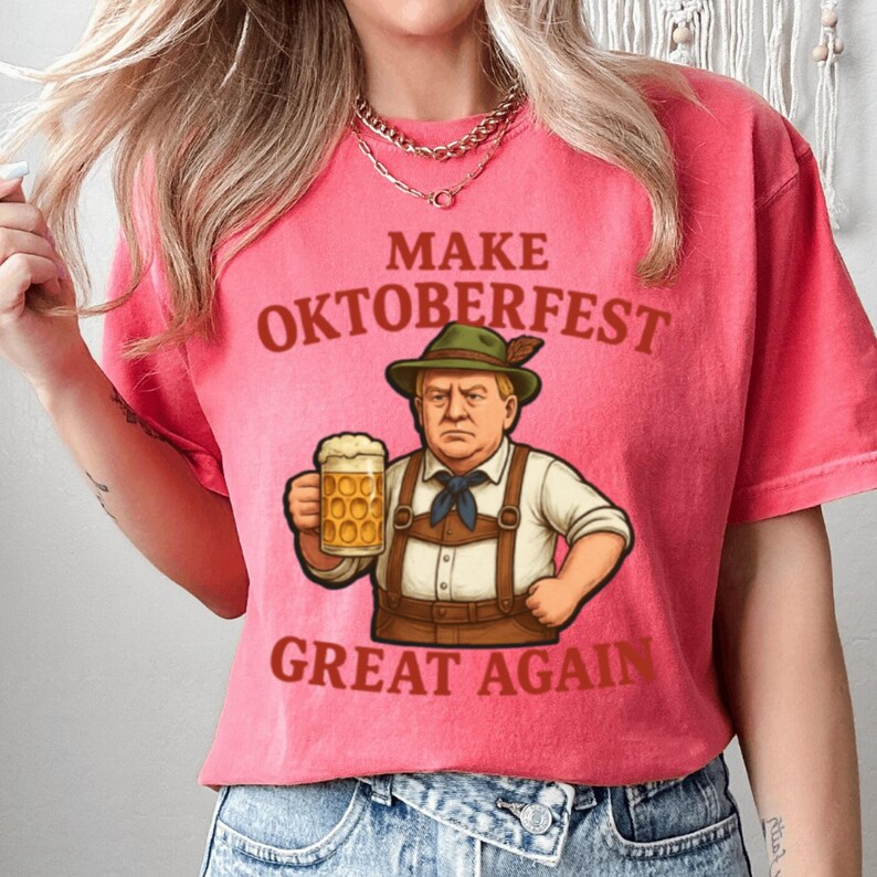 May include: Pink t-shirt with the text "MAKE OKTOBERFEST GREAT AGAIN" and a cartoon illustration of a man in traditional Bavarian attire holding a beer stein. The design features brown and green colors.