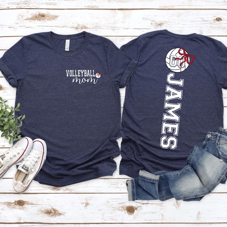 May include: Navy blue t-shirts with volleyball-themed designs. One shirt says "Volleyball Mom" with a volleyball and bow graphic. The other shirt has the name "JAMES" vertically with a volleyball and bow graphic. White sneakers and blue jeans are also visible.