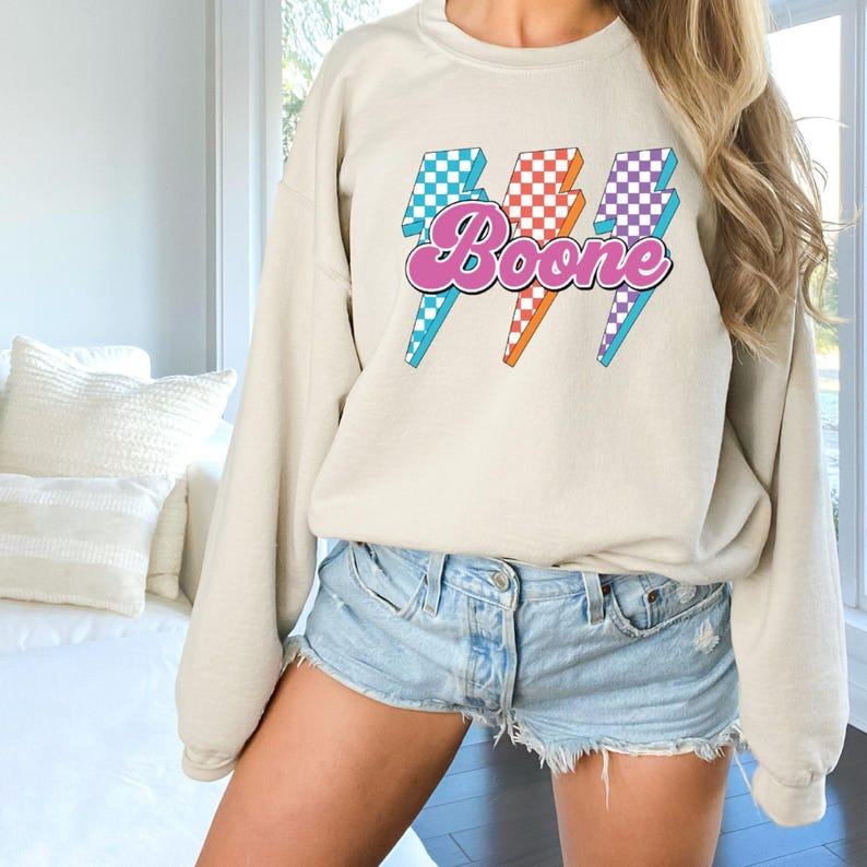 May include: A beige sweatshirt with the name "Boone" in a pink, retro font, overlaid on three lightning bolt graphics. The lightning bolts feature checkered patterns in teal, orange, and purple. The sweatshirt has long sleeves and a crew neck.