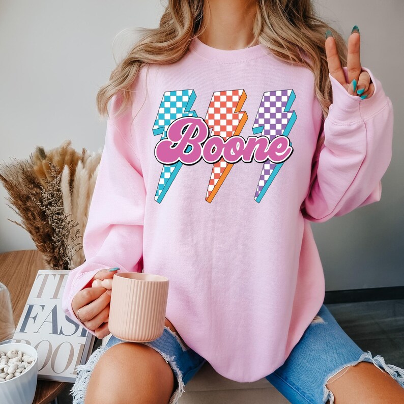 May include: A light pink sweatshirt with the name "Boone" in a retro font, outlined in pink, with three lightning bolt graphics. The lightning bolts have checkered patterns in blue, orange, and purple. The sweatshirt is a casual style.