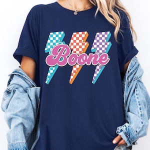 May include: Navy blue t-shirt featuring the name "Boone" in a retro font, overlaid on three lightning bolt graphics. The lightning bolts have checkered patterns in blue, red, and purple, with orange and white accents.