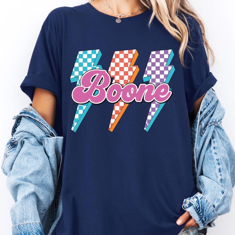 May include: Navy blue t-shirt featuring the name "Boone" in a retro font, overlaid on three lightning bolt graphics. The lightning bolts have checkered patterns in blue, red, and purple, with orange and white accents.