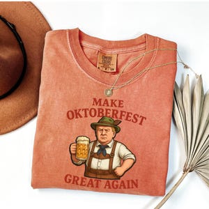 May include: Coral-colored Comfort Colors t-shirt with the text "MAKE OKTOBERFEST GREAT AGAIN" and a cartoon image of a man in traditional Bavarian attire holding a beer stein. A gold necklace and a brown hat are also visible.