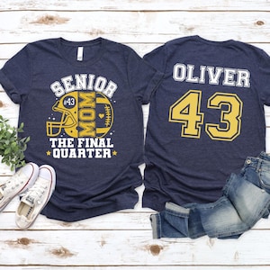 May include: A navy blue t-shirt featuring a yellow football helmet graphic with the text SENIOR MOM THE FINAL QUARTER and the number 43. The back of the shirt displays the name OLIVER in white letters above the large yellow number 43.