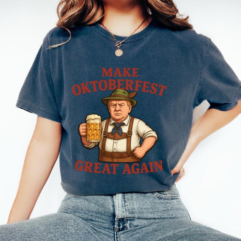 May include: Navy blue t-shirt with the phrase "MAKE OKTOBERFEST GREAT AGAIN" in red text. The graphic features a cartoon of a man in traditional Bavarian attire holding a beer stein. The shirt is a comfortable, casual style.