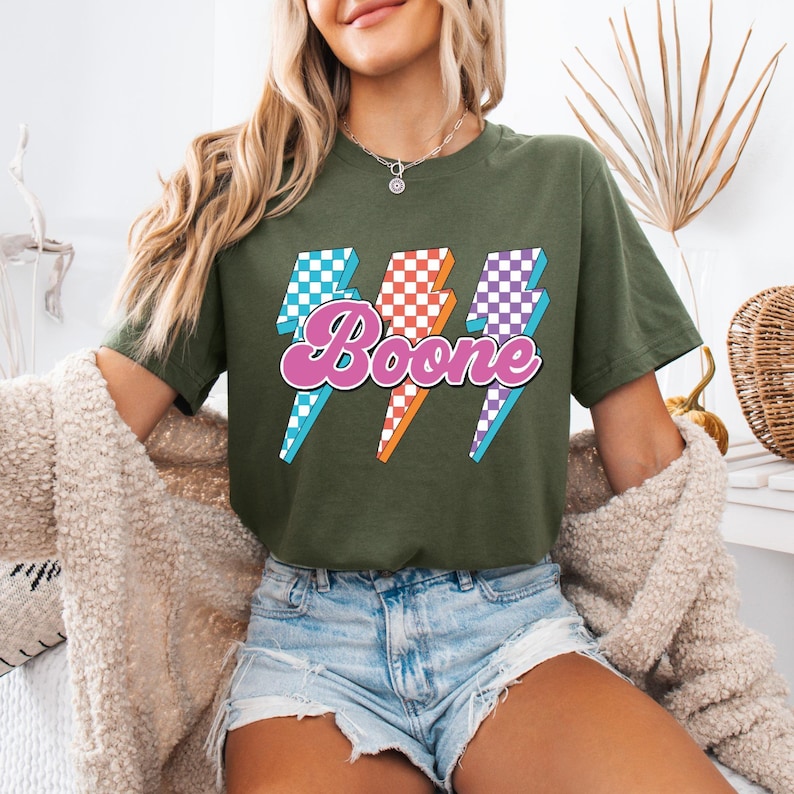May include: Olive green t-shirt featuring the name "Boone" in a pink and purple script font, with three lightning bolt graphics in blue, orange, and purple checkered patterns. The shirt is a casual style.