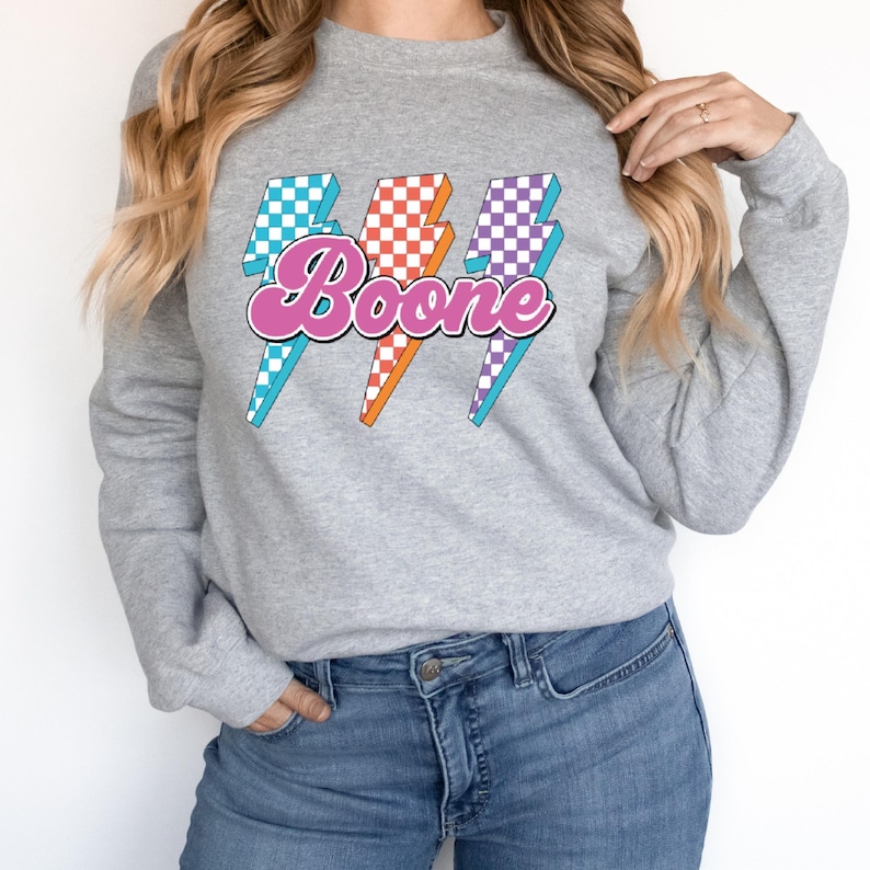 May include: A gray sweatshirt featuring three lightning bolt graphics with a checkered pattern in blue, orange, and purple. The word "Boone" is written in pink script across the center of the lightning bolts.