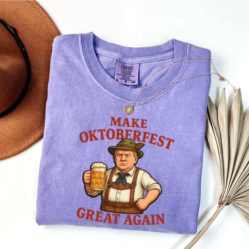 May include: A lilac Comfort Colors t-shirt with the phrase "MAKE OKTOBERFEST GREAT AGAIN" in red lettering. The graphic features a cartoon of a man in traditional Bavarian attire holding a beer stein. A gold necklace is draped across the shirt.