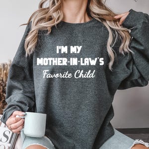 May include: Dark gray sweatshirt with the text "I'M MY MOTHER-IN-LAW'S Favorite Child" in white. The sweatshirt is a crew neck style and appears to be made of a soft material. The text is centered on the front of the shirt.