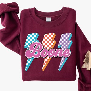 May include: A maroon sweatshirt with the name "Boone" in a pink, retro font, overlaid on three lightning bolt graphics. The lightning bolts have a checkered pattern in blue and white, orange and white, and purple and white.