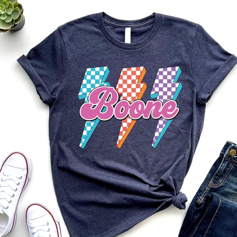 May include: Navy blue t-shirt with a retro design featuring the name "Boone" in a pink script font, overlaid on three lightning bolt graphics. The lightning bolts have checkered patterns in blue, orange, and purple.