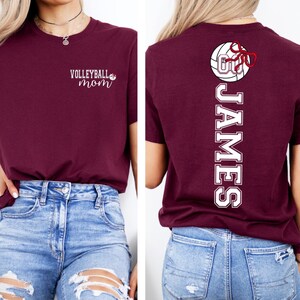 May include: Maroon t-shirt with the text "VOLLEYBALL mom" on the front. The back of the shirt features a volleyball graphic with a red bow and the name "JAMES" in white lettering. The shirt is paired with blue jeans.
