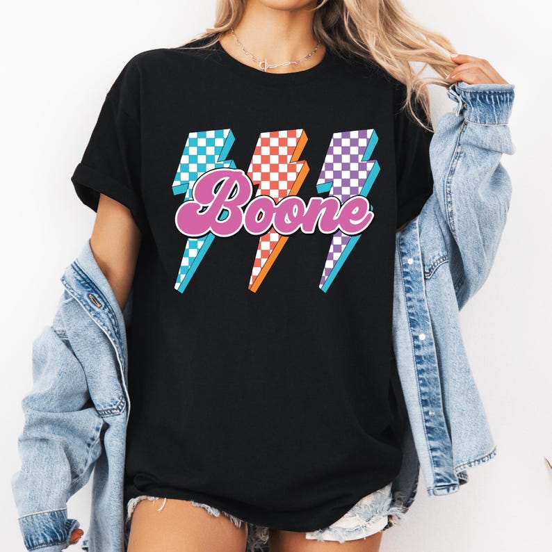 May include: Black t-shirt featuring the name "Boone" in a pink, retro font, with three lightning bolt graphics in blue, orange, and purple checkered patterns. The shirt is a casual, everyday wear item.