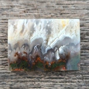 Peut inclure: Une pierre rectangulaire avec un motif naturel et abstrait. Le motif présente des teintes blanches, grises, marron, vertes et orange, ressemblant à un paysage ou à une formation nuageuse. La pierre est posée sur une surface en bois.