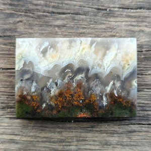 Könnte beinhalten: Ein rechteckiger Stein mit Landschaftsdesign. Der obere Teil ist weiß und gelb, geht in der Mitte in Grau und Braun über und unten in Grün und Orange. Der Stein liegt auf einer Holzoberfläche.