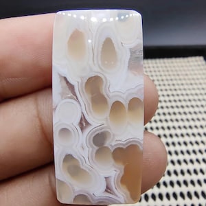 May include: A rectangular gemstone with a unique pattern of swirling white, tan, and brown colors. The polished surface reveals a natural, organic design, perfect for jewelry making or collecting.